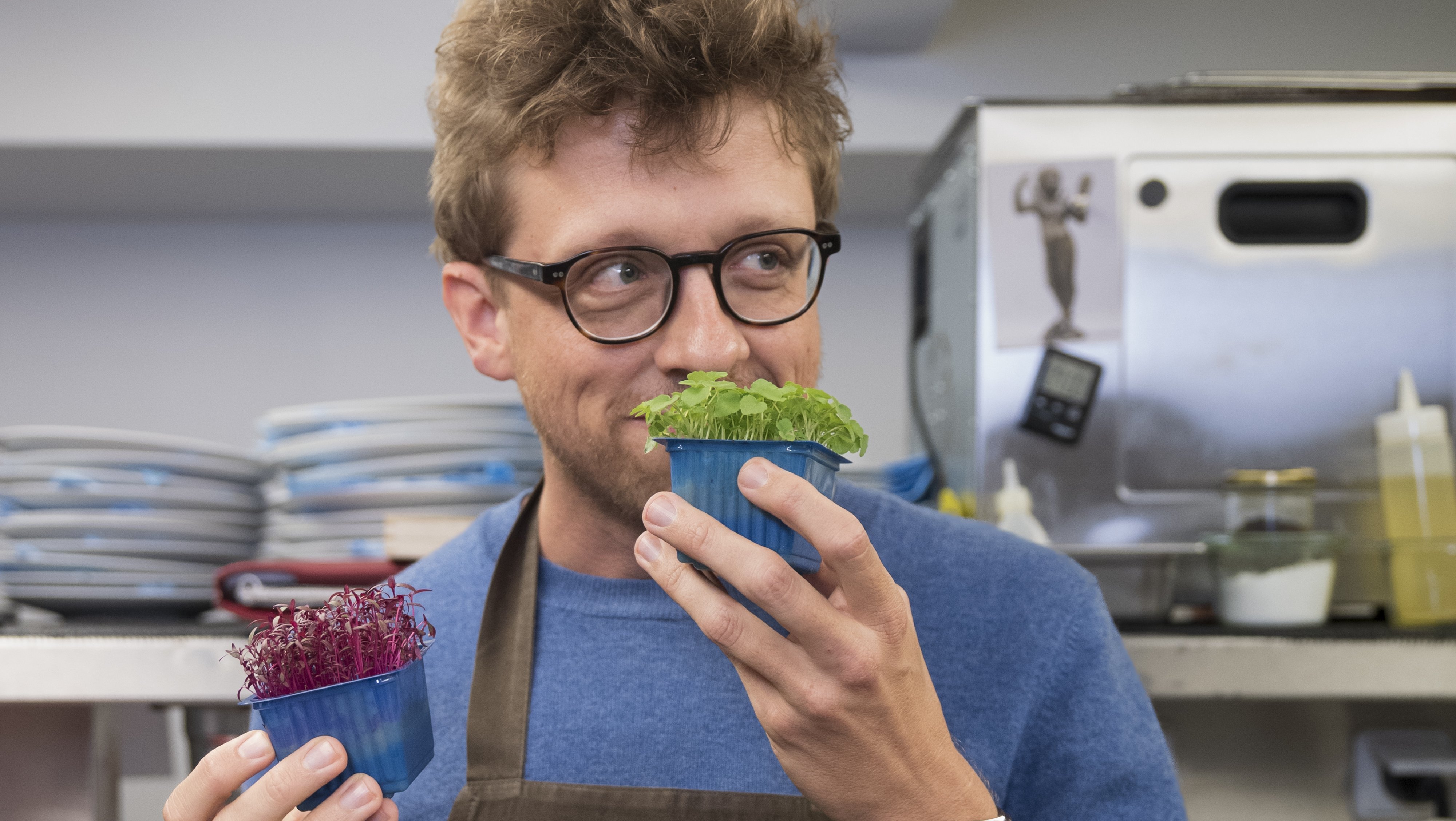 Martin von der Nahmer is standing in a kitchen while holding a green herb plant to his nose. In his other hand, he is holding a red coloured plant.