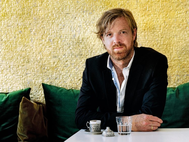 Matthew McDonald is sitting on a green sofa infront of a white table. 