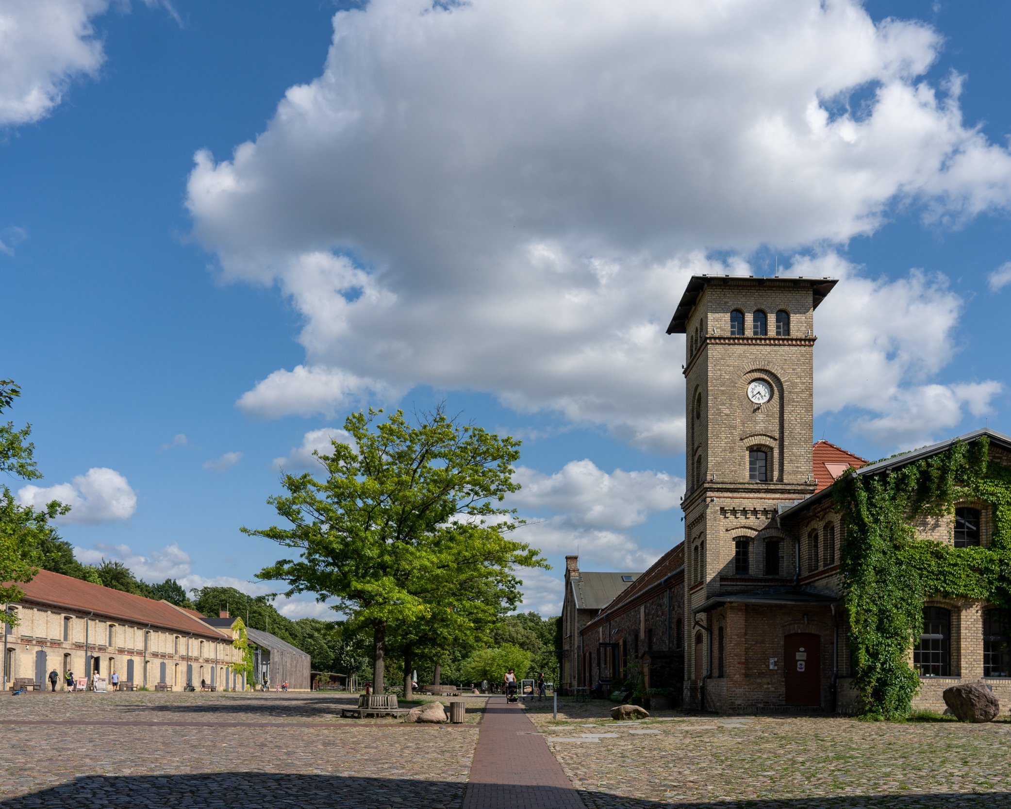 Historischer Gutshof mit Kopfsteinpflaster, einem Turm mit Uhr und Gebäuden mit roten Dächern sowie begrünten Fassaden unter einem blauen Himmel mit Wolken.