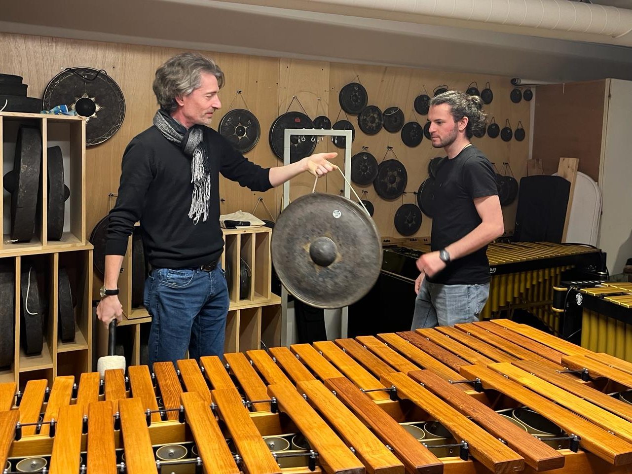 Two men stand in a room full of percussion instruments, including gongs and marimbas. One is holding a gong and a mallet while the other looks on.