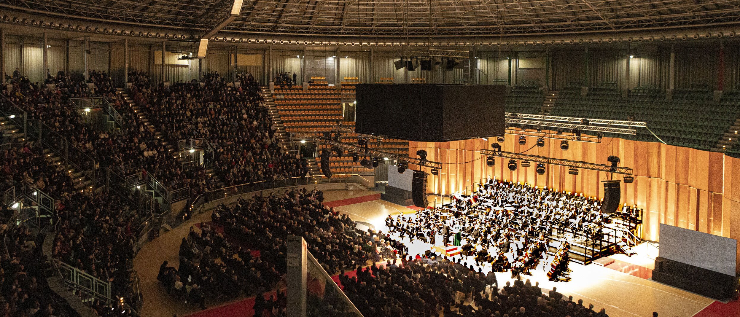 A large orchestra plays in a modern, semi-circular concert hall in front of a full audience under an impressive domed roof.