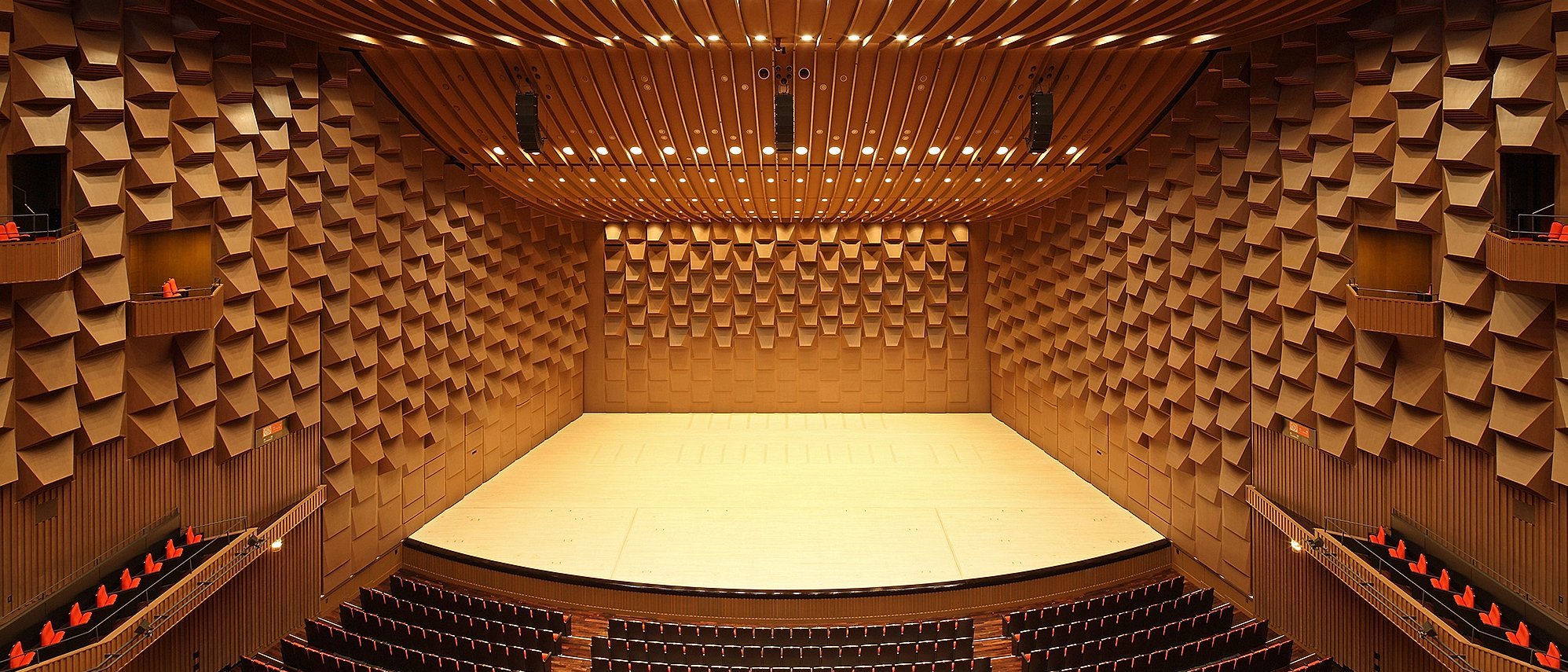 A concert hall with a view of the stage, the walls are wood-panelled and the seats are covered in red.