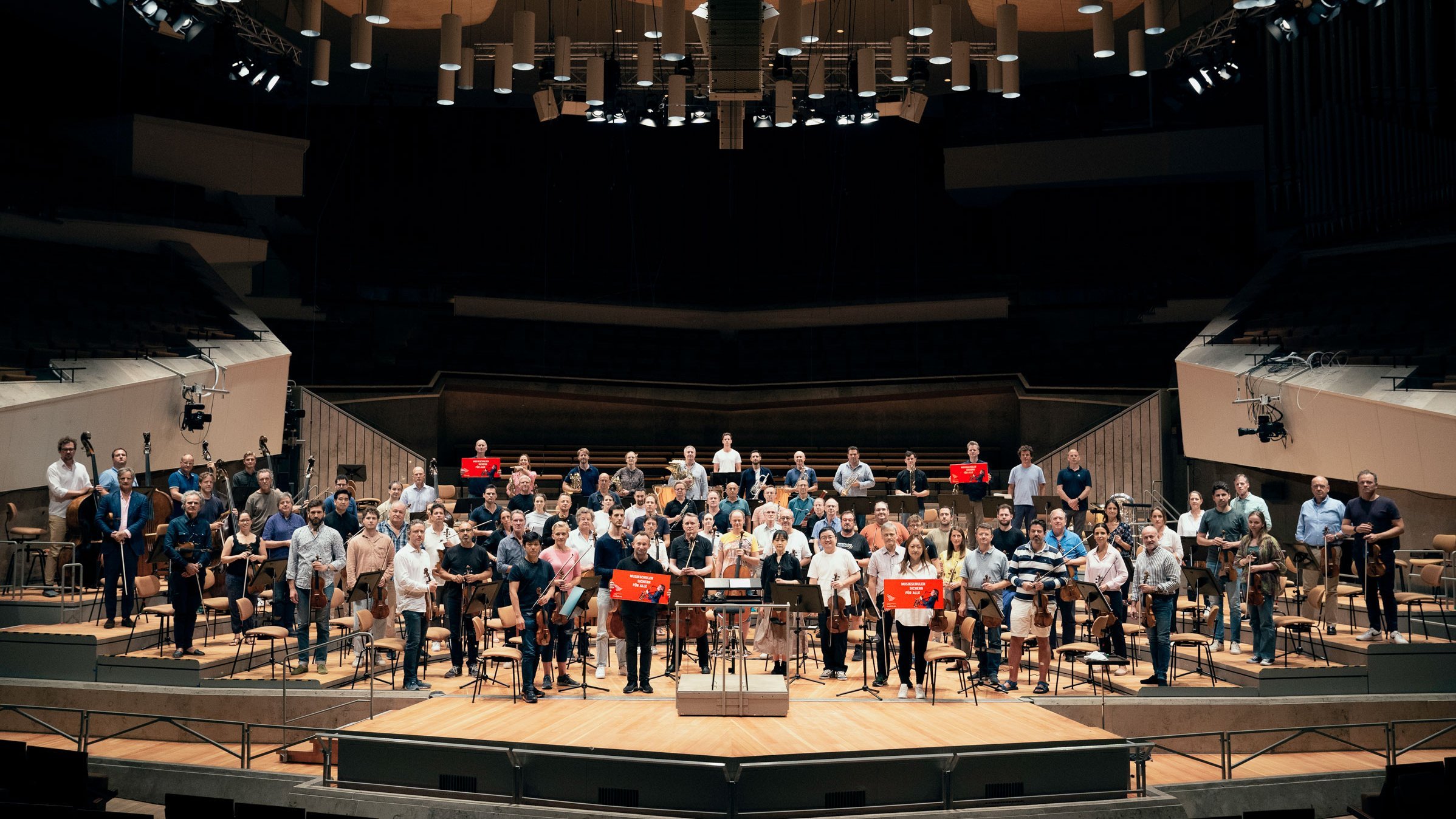 Die Berliner Philharmoniker und Kirill Petrenko stehen auf der Bühne der Philharmonie und halten Plakta hoch. Darauf steht »Musikschule sichern für alle!«