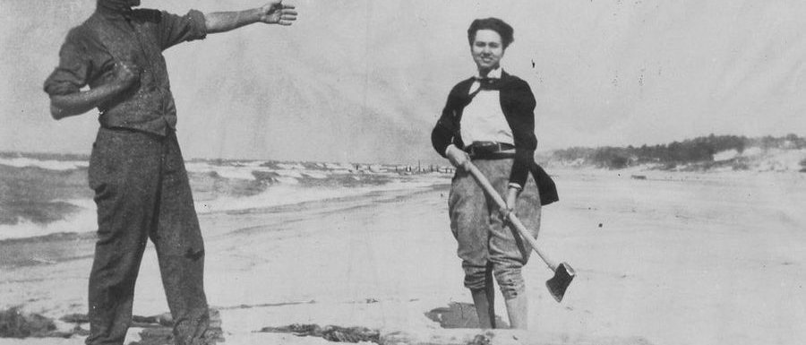 Ein Mann deutet auf eine Frau, die am Strand mit Beil über einem Baumstamm steht