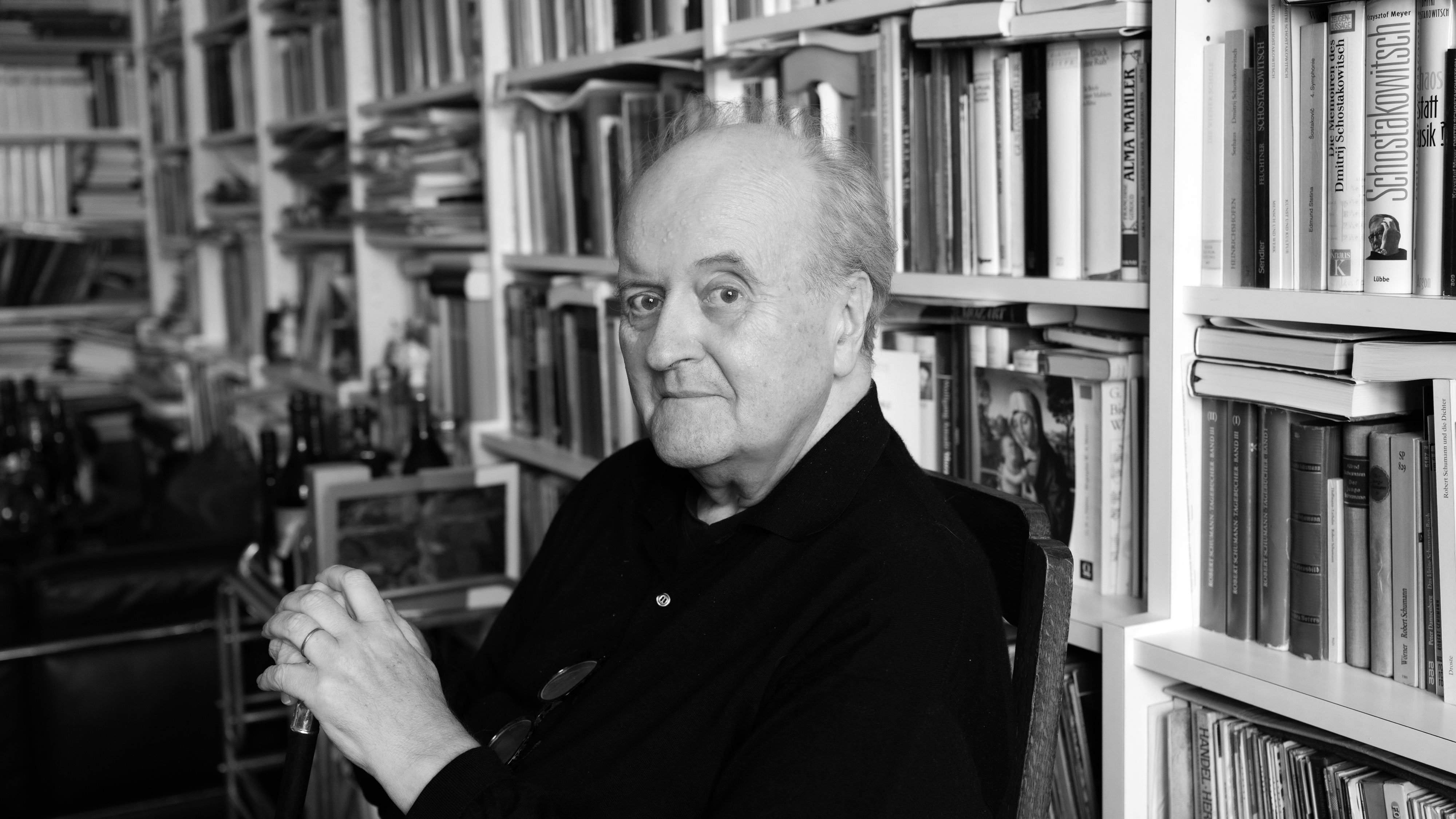 An older man is sitting in front of a full bookshelf. He has thinning hair and is wearing a dark jumper. He is holding a walking stick in his hand. The picture is in black and white.