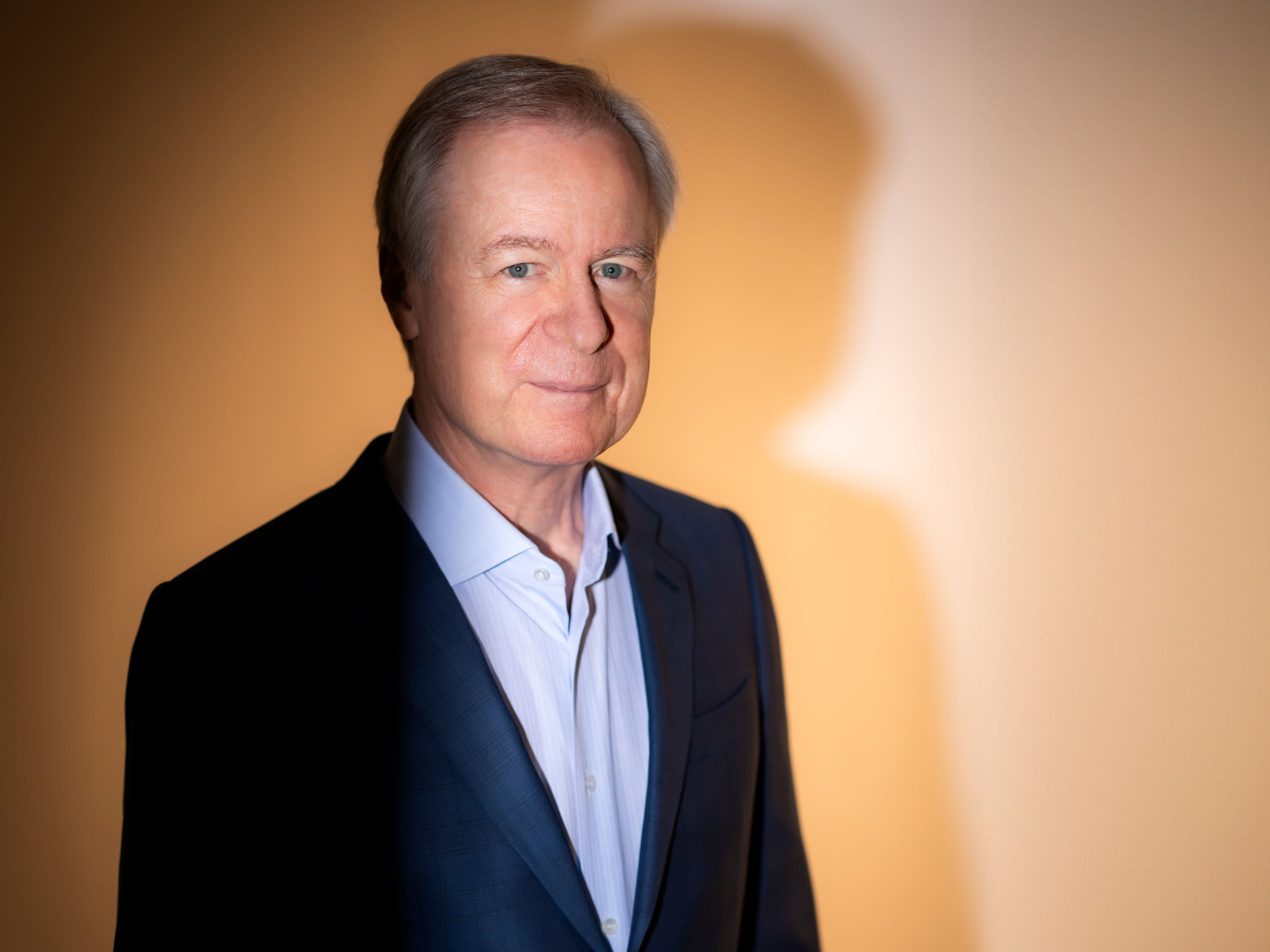 A person in a blue suit and with grey hair stands in front of an orange-yellow illuminated wall. A clear shadow can be seen on it.