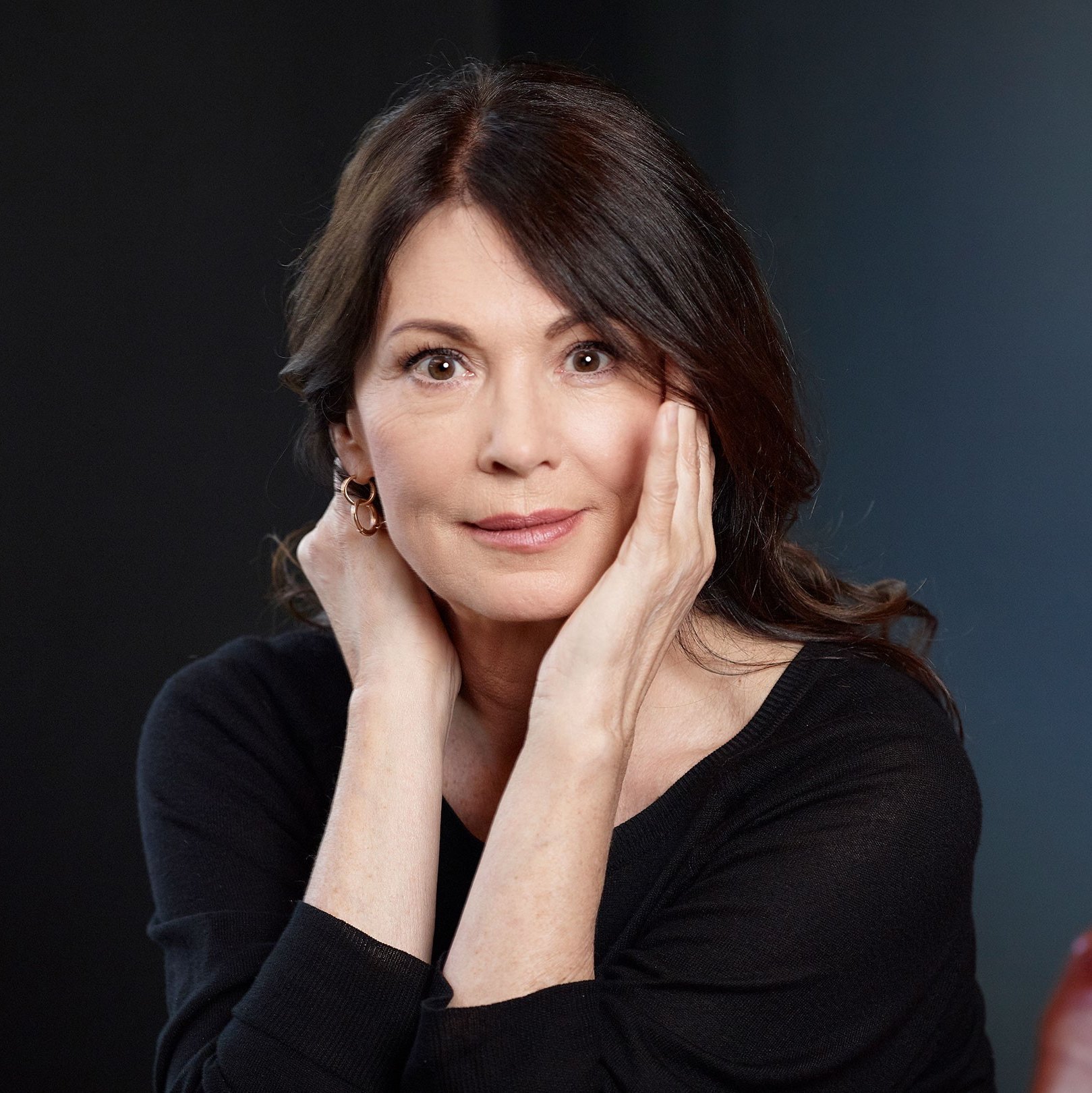 Iris Berben is wearing a black sweater, her elbows are propped up and her hands are on her face. The background is dark blue.