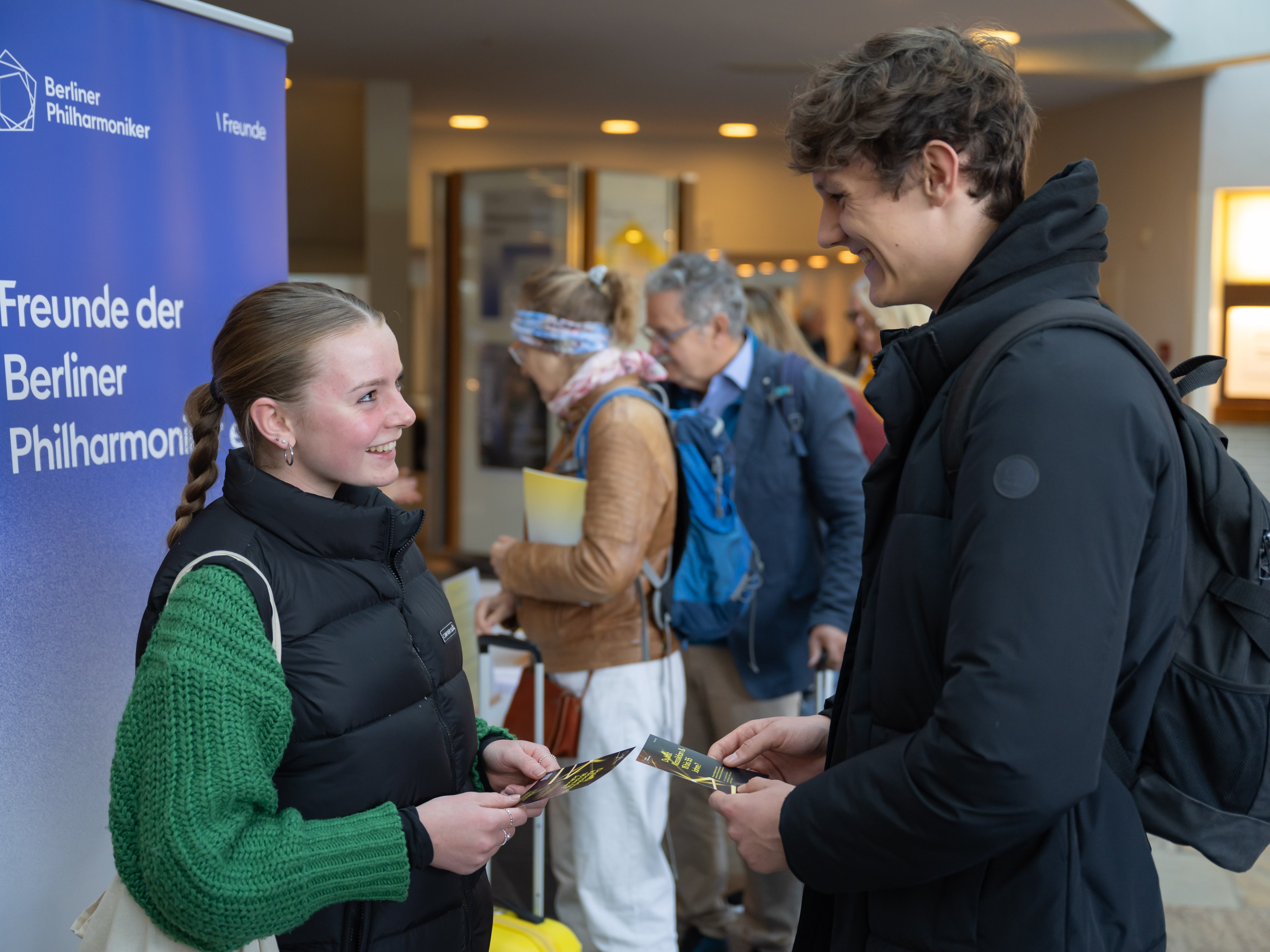 Several people are standing in the foyer of the Berlin Philharmonie, chatting. Some are holding programme booklets. The background shows other guests and architectural details of the foyer.