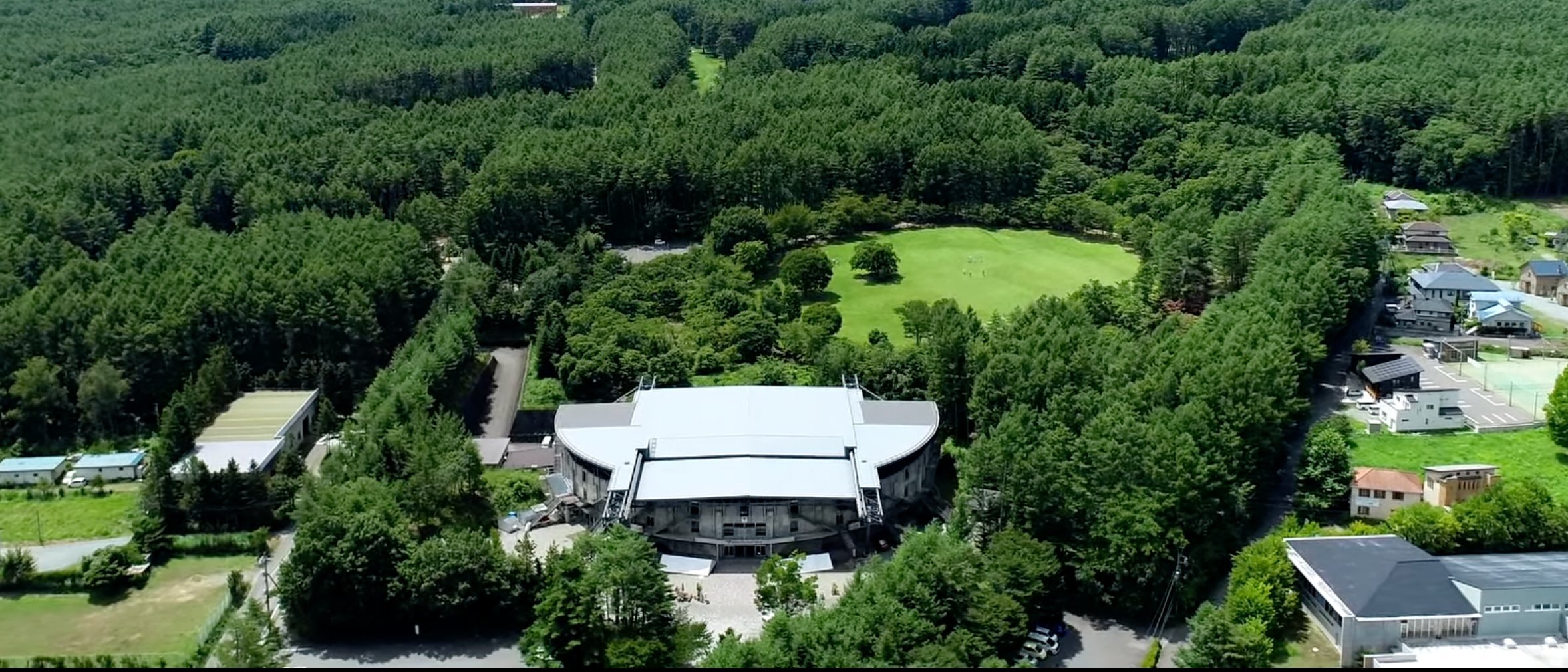 A modern theatre surrounded by a few houses and lots of forest in the town of Kawaguchiko. Mount Fuji can be seen on the horizon.