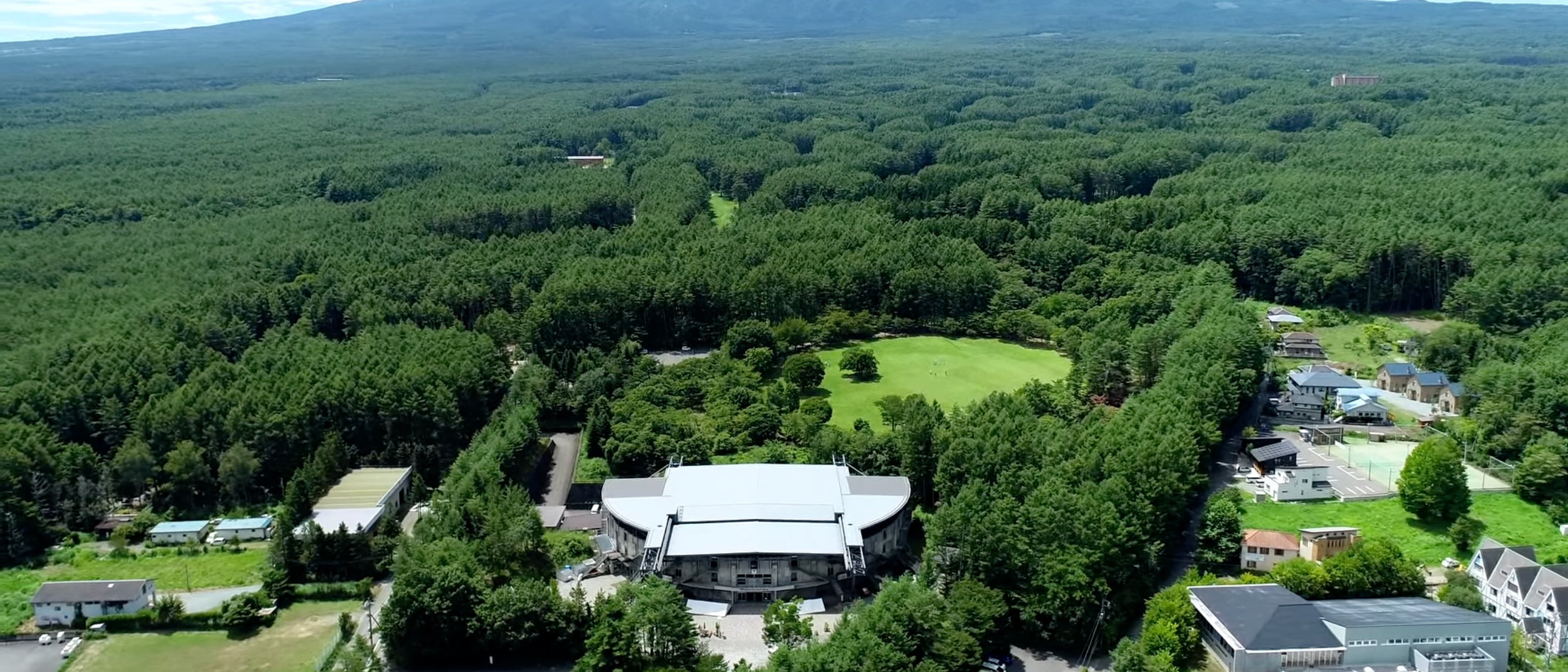 A modern theatre surrounded by a few houses and lots of forest in the town of Kawaguchiko. Mount Fuji can be seen on the horizon.