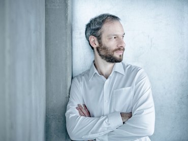 Einer Person mit kurzen grau-schwarzen Haaren und Bart lehnt mit verschränkten Armen an einer Betonwand. Die Person schaut nach rechts und trägt ein weißes Hemd.