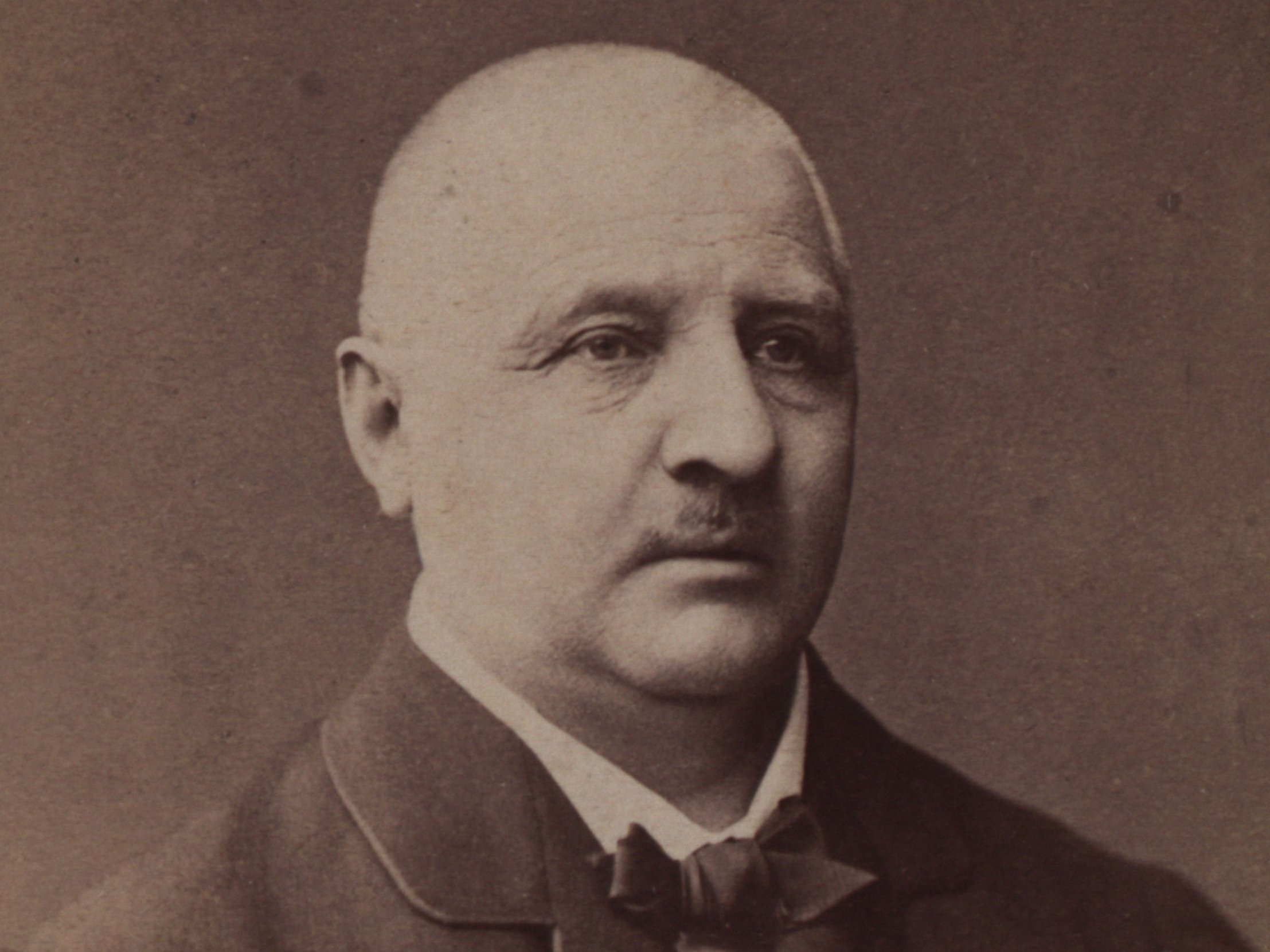 Photograph in shades of brown of Anton Bruckner, he looks past the camera, his jacket is a bit big, he wears a bow tie, has only little hair and a short moustache