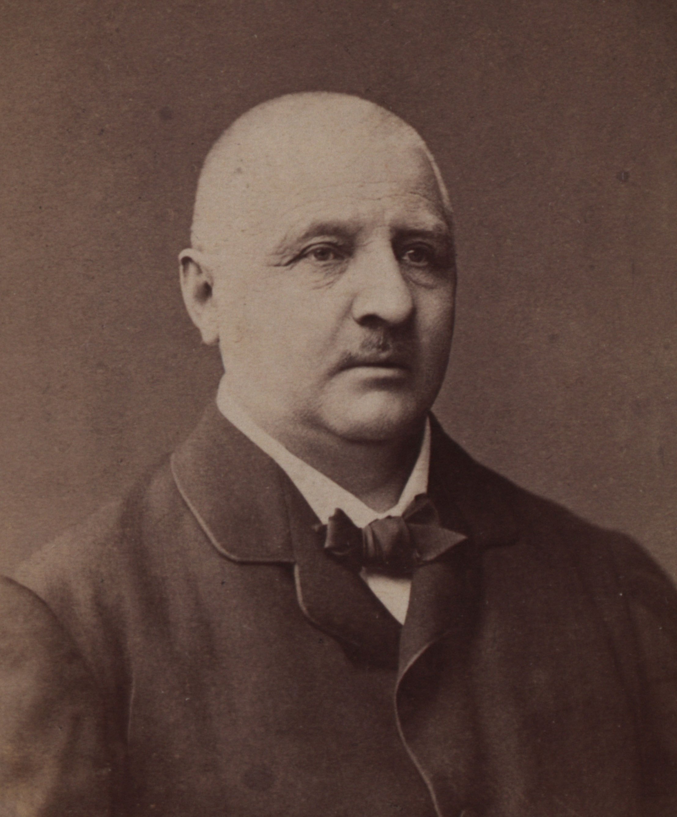 Photograph in shades of brown of Anton Bruckner, he looks past the camera, his jacket is a bit big, he wears a bow tie, has only little hair and a short moustache