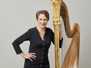 Marie-Pierre Langlamet stands in front of a grey background, holding her harp with her left hand and her right on her hip.