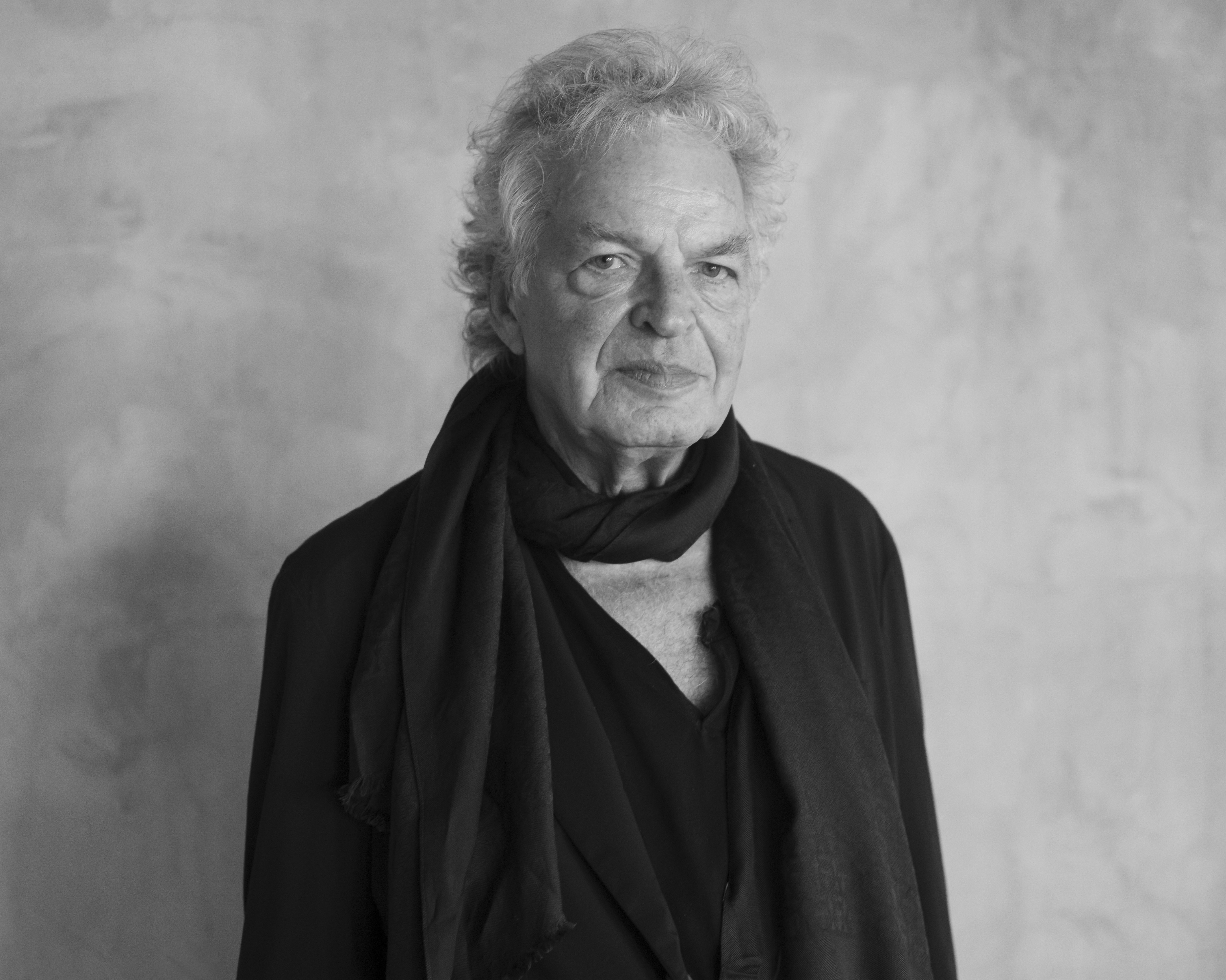 A black and white photo of a man with white hair. He is wearing a black shirt and jacket and a black scarf wrapped around his neck. 