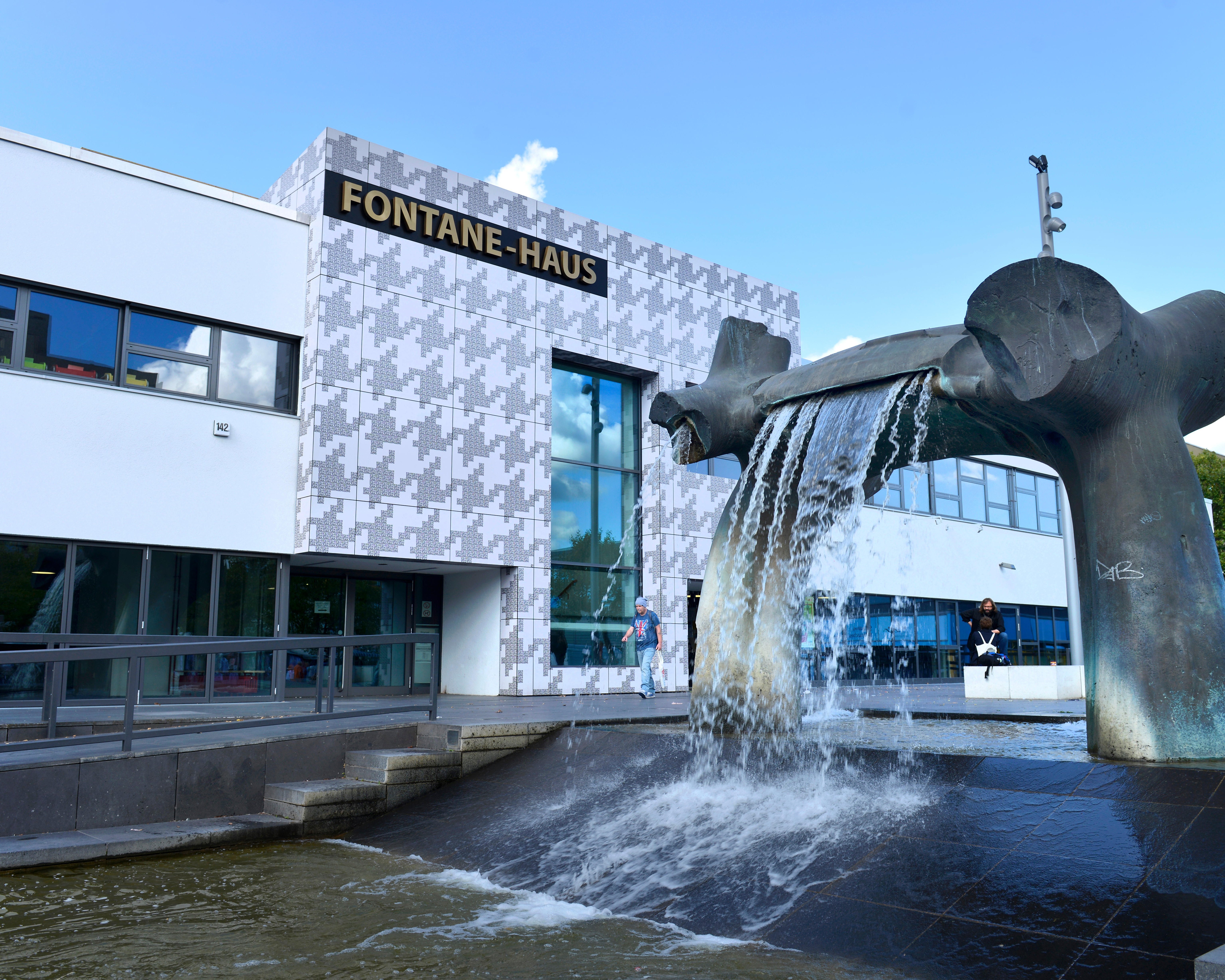 Das Bild zeigt das Fontane-Haus in Berlin, ein modernes Gebäude mit weißer Fassade und Musterung. Im Vordergrund befindet sich ein großer, skulpturaler Brunnen mit Wasserfällen, der die Szene dynamisch belebt. 