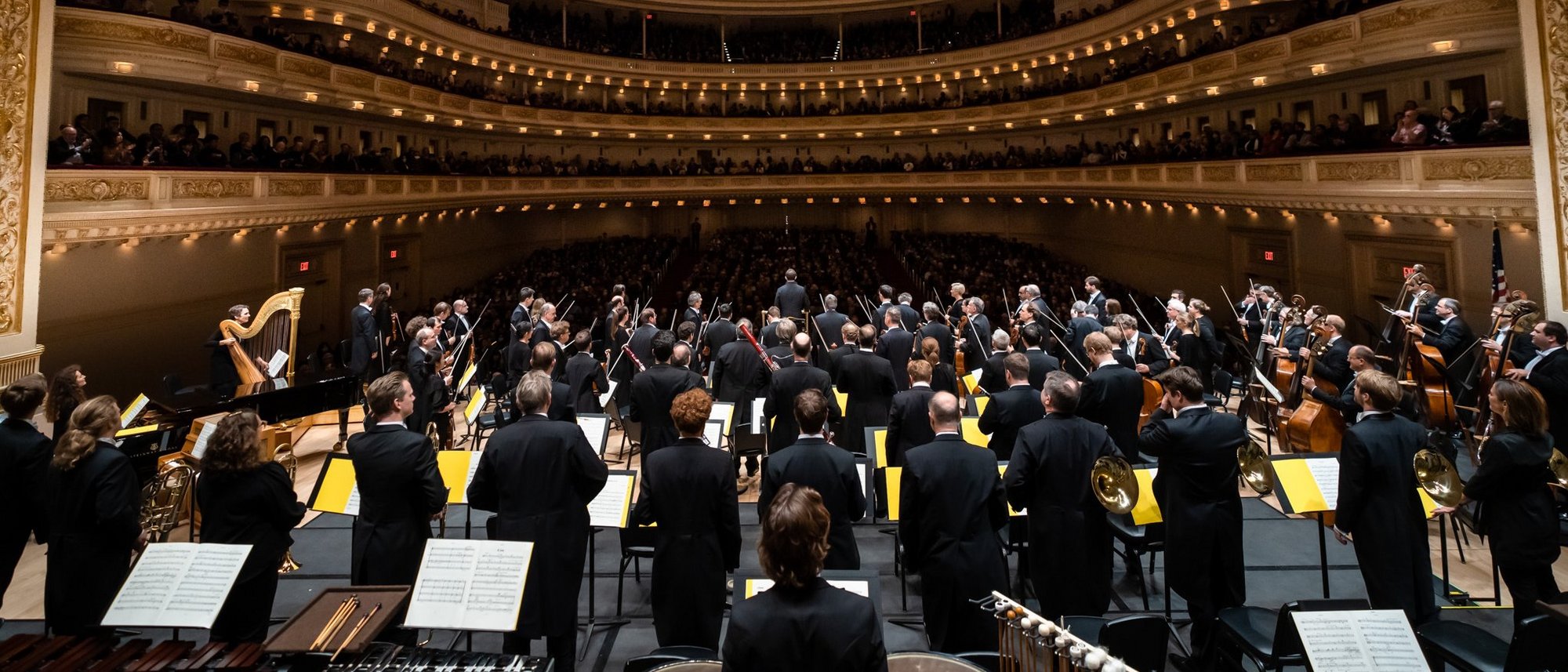 View behind the orchestra towards the audience in Carnegie Hall. The orchestra stands.