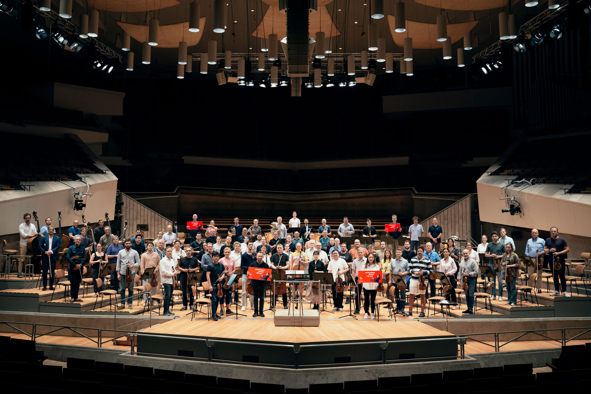 Die Berliner Philharmoniker und Kirill Petrenko stehen auf der Bühne der Philharmonie und halten Plakta hoch. Darauf steht »Musikschule sichern für alle!«