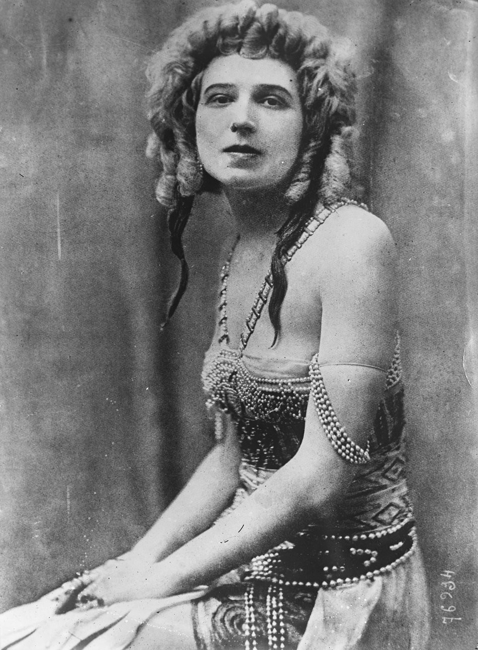 Das Bild zeigt eine historische Schwarz-Weiß-Fotografie von Ida Rubinstein in einem aufwendig verzierten Bühnenkostüm mit Perlen. Sie sitzt mit gefalteten Händen und voluminösem lockigem Haar vor einem neutralen Hintergrund.