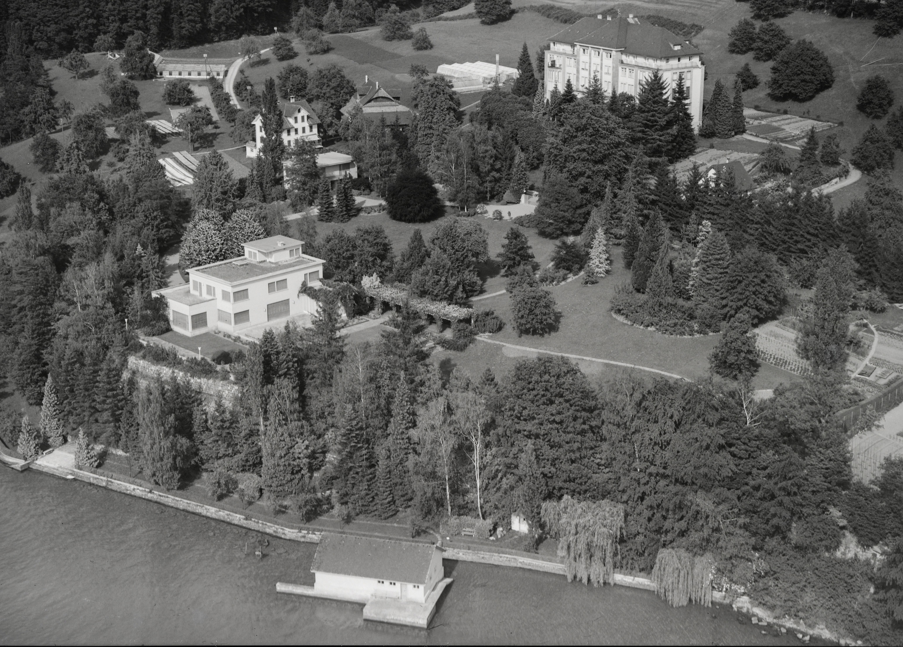 Aerial view of a spacious estate on the shore of a lake. A modern villa can be seen in the foreground, a boathouse by the water and a park-like landscape with numerous trees, paths and gardens. In the background is a large building that looks like a manor house or hotel, embedded in a rural setting.