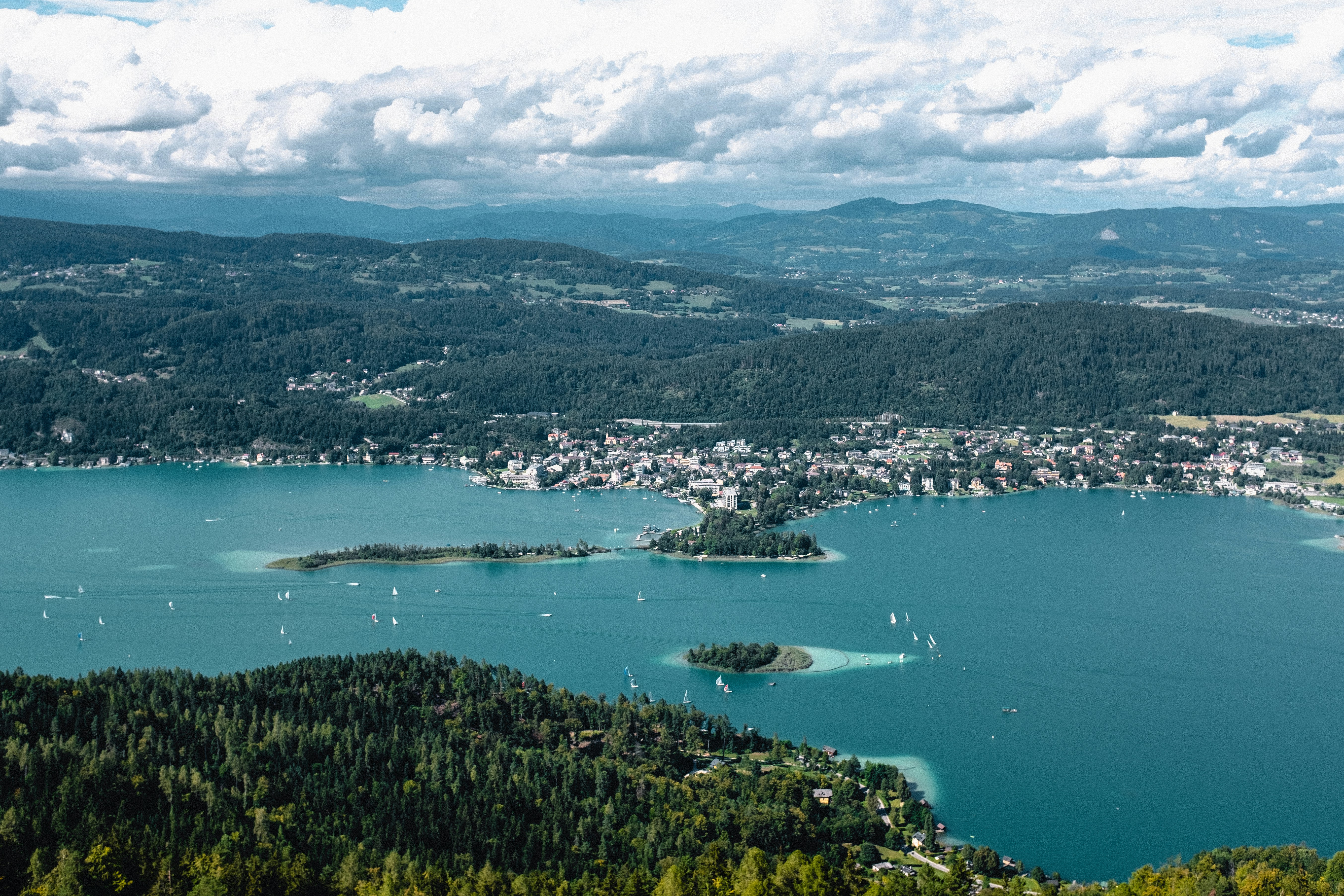 Bird’s-eye view of the lake and shore, the water is blue-green, sailing boats are sailing on the lake, there are a few clouds in the sky