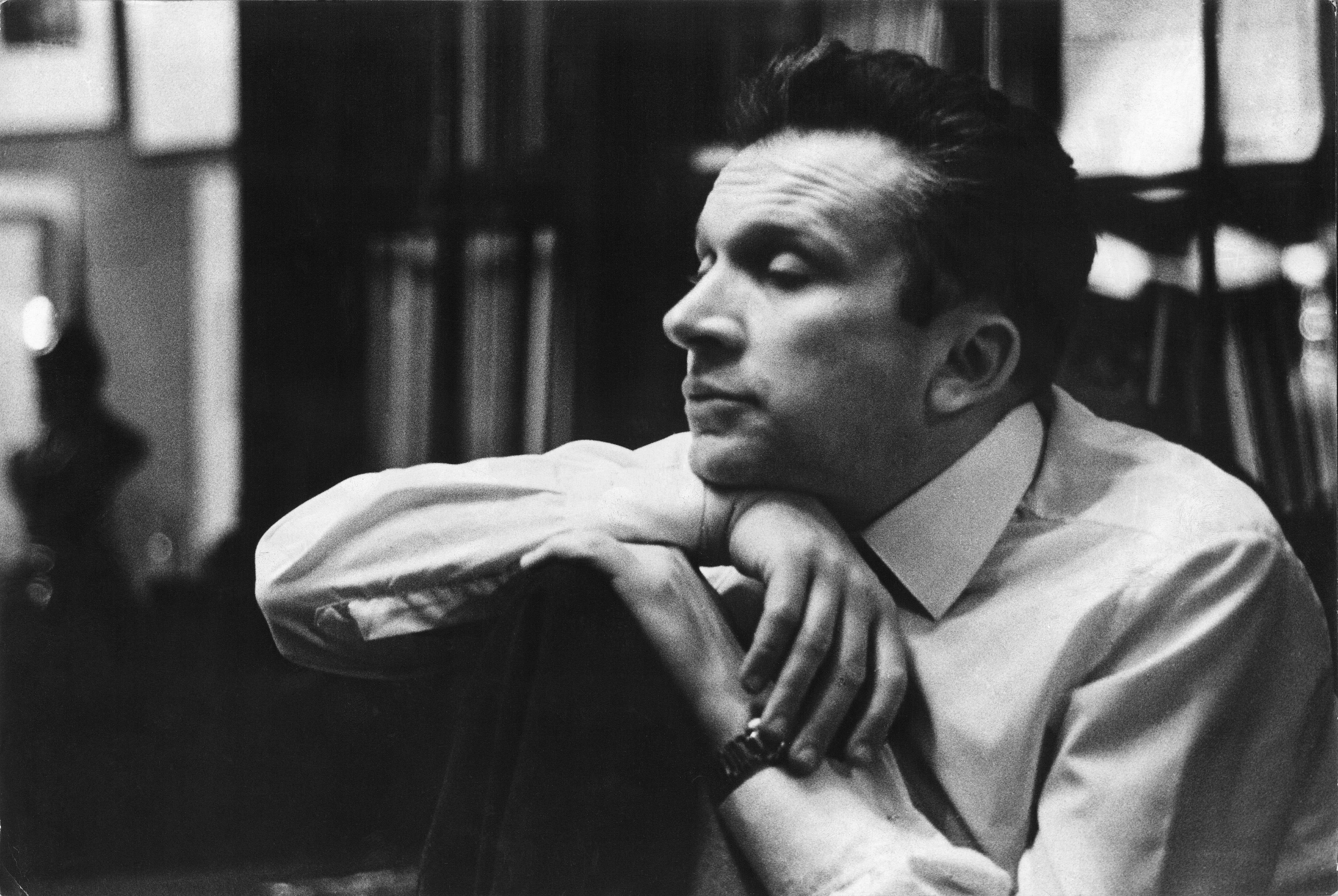 The picture shows the composer Mieczysław Weinberg looking thoughtfully to the side. His chin rests on his folded arms, which are resting on the back of a chair. He is wearing a white shirt .