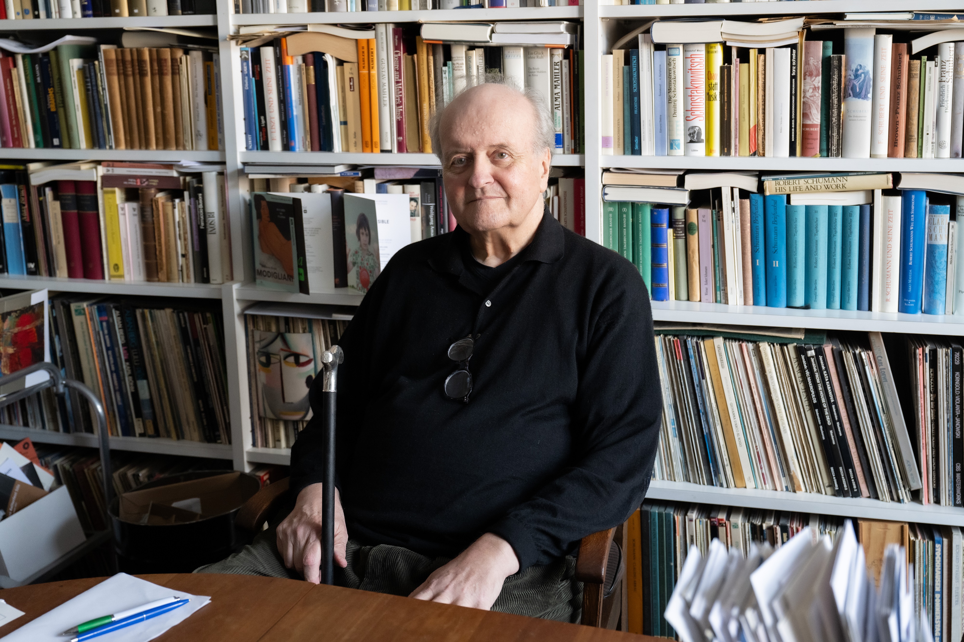 An older man is sitting in front of a full bookshelf. He has thinning hair and is wearing a dark jumper. He is holding a walking stick in his hand. The picture is in black and white.