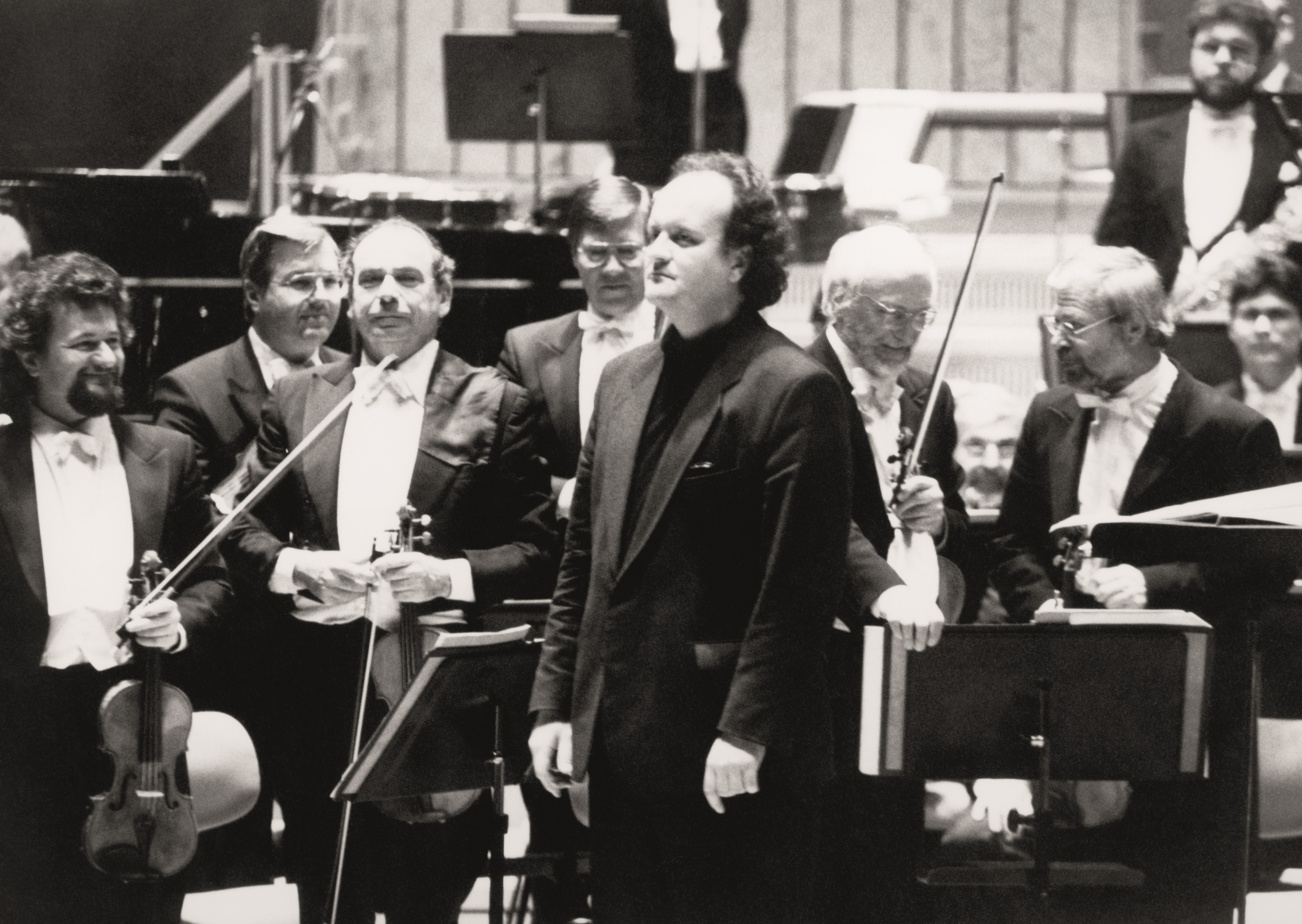 Wolfgang Rihm in a suit stands in front of the orchestra, the musicians stand and hold their instruments in their hands.