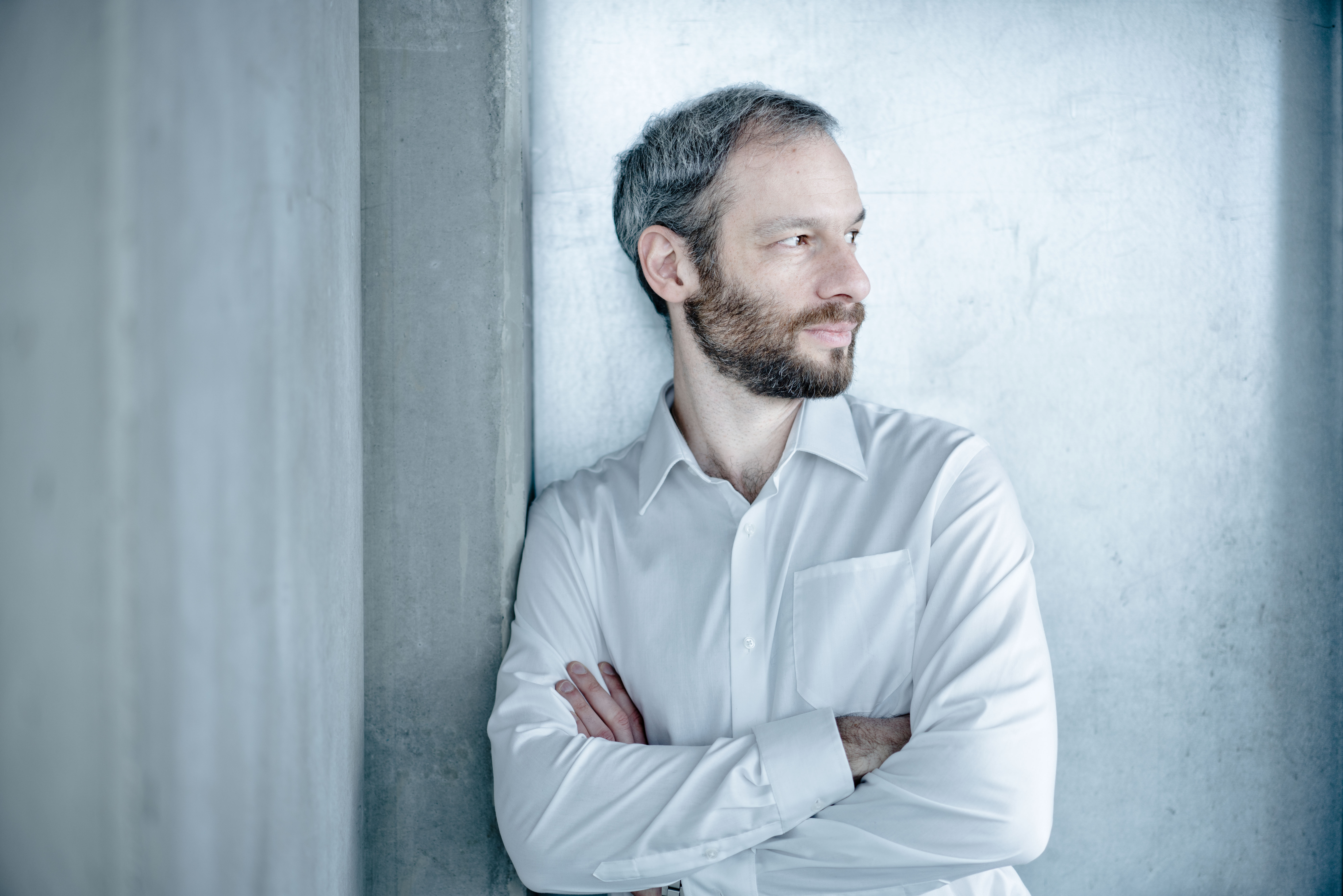 A person with short grey-black hair and a beard is leaning against a concrete wall with his arms crossed. The person is looking to the right and is wearing a white shirt.