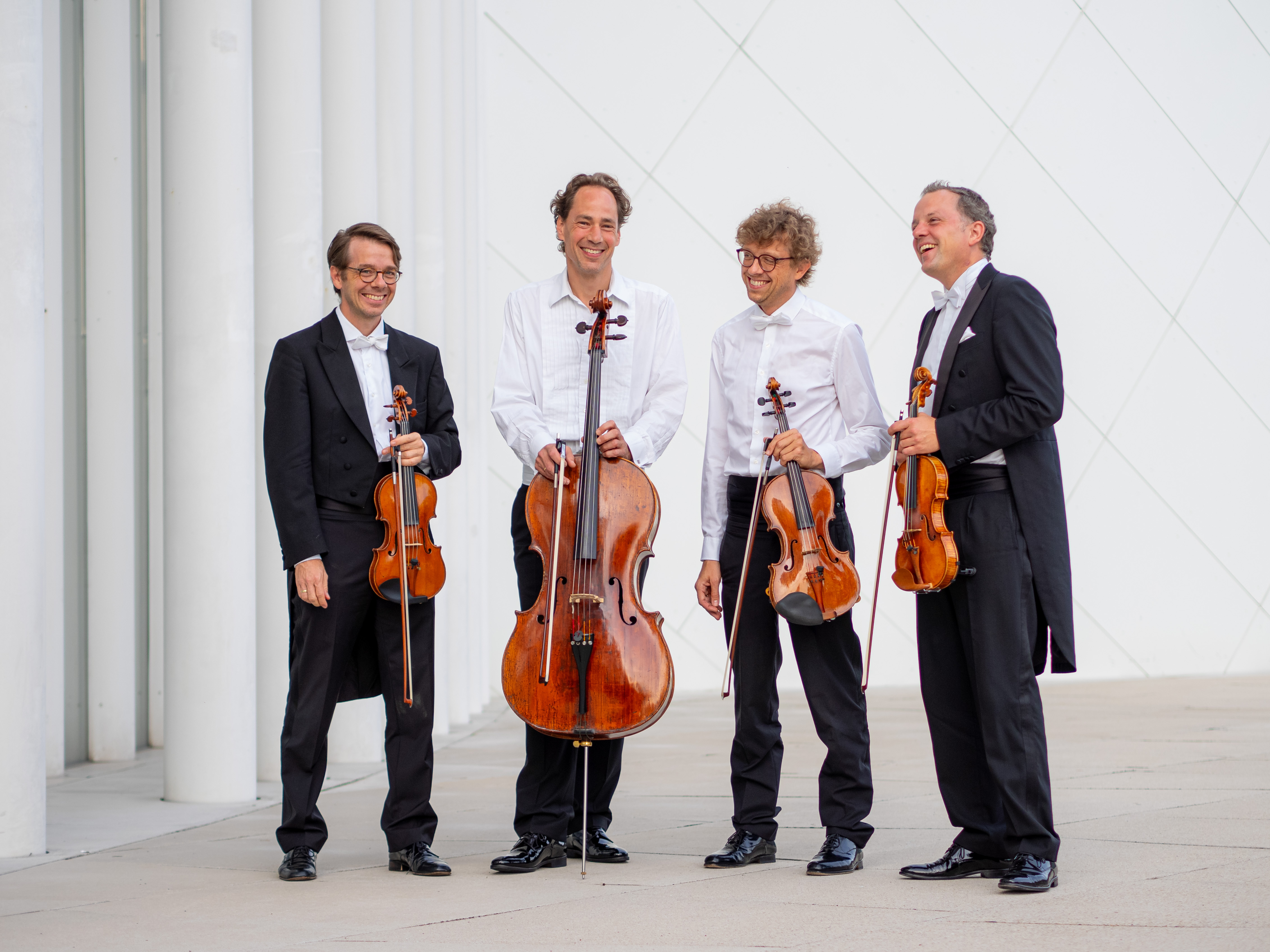 Four male musicians stand in tails, some in white shirts on white pillars. They are holding their instruments, two violins, a viola and a cello.