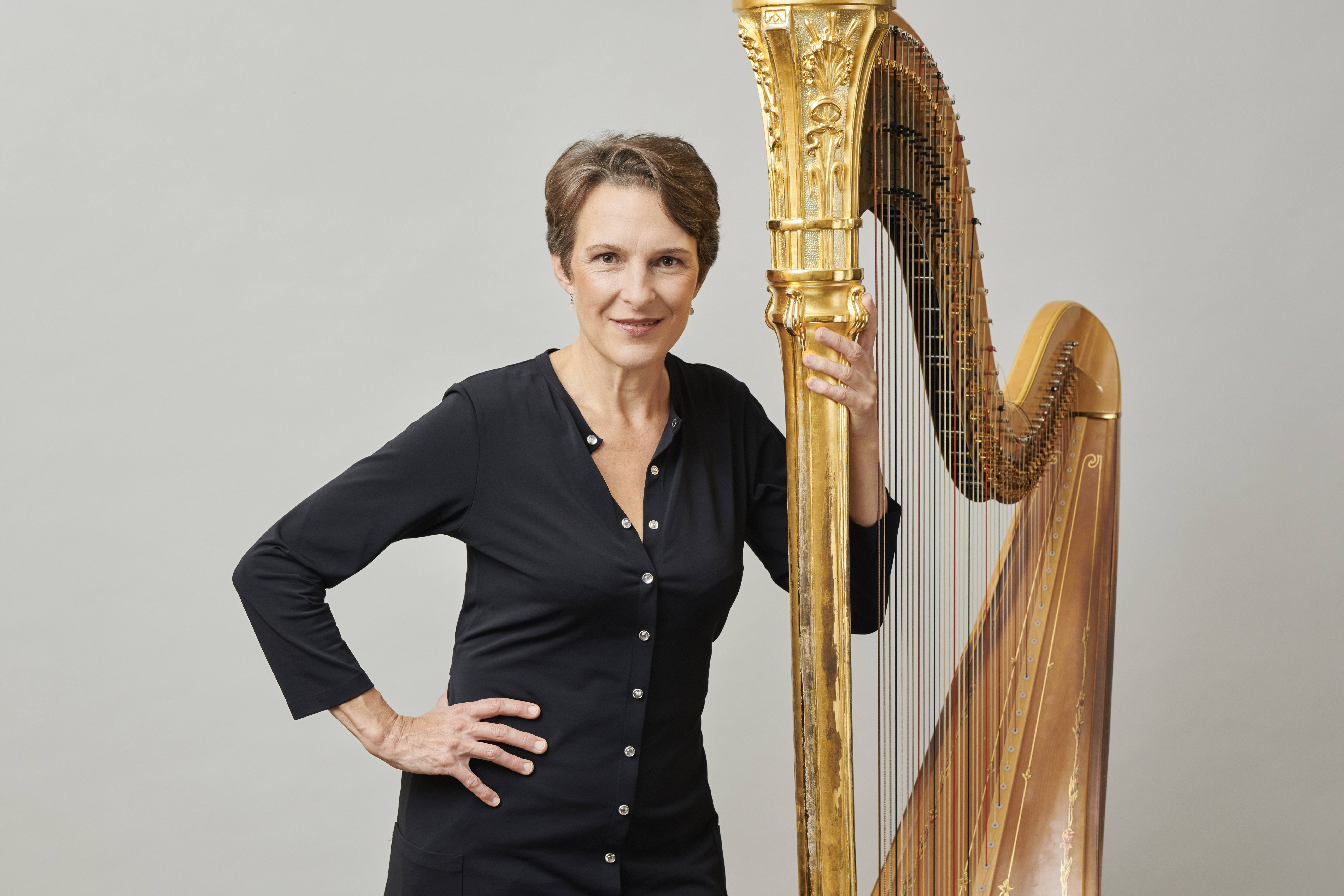 Marie-Pierre Langlamet stands in front of a grey background, holding her harp with her left hand and her right on her hip.