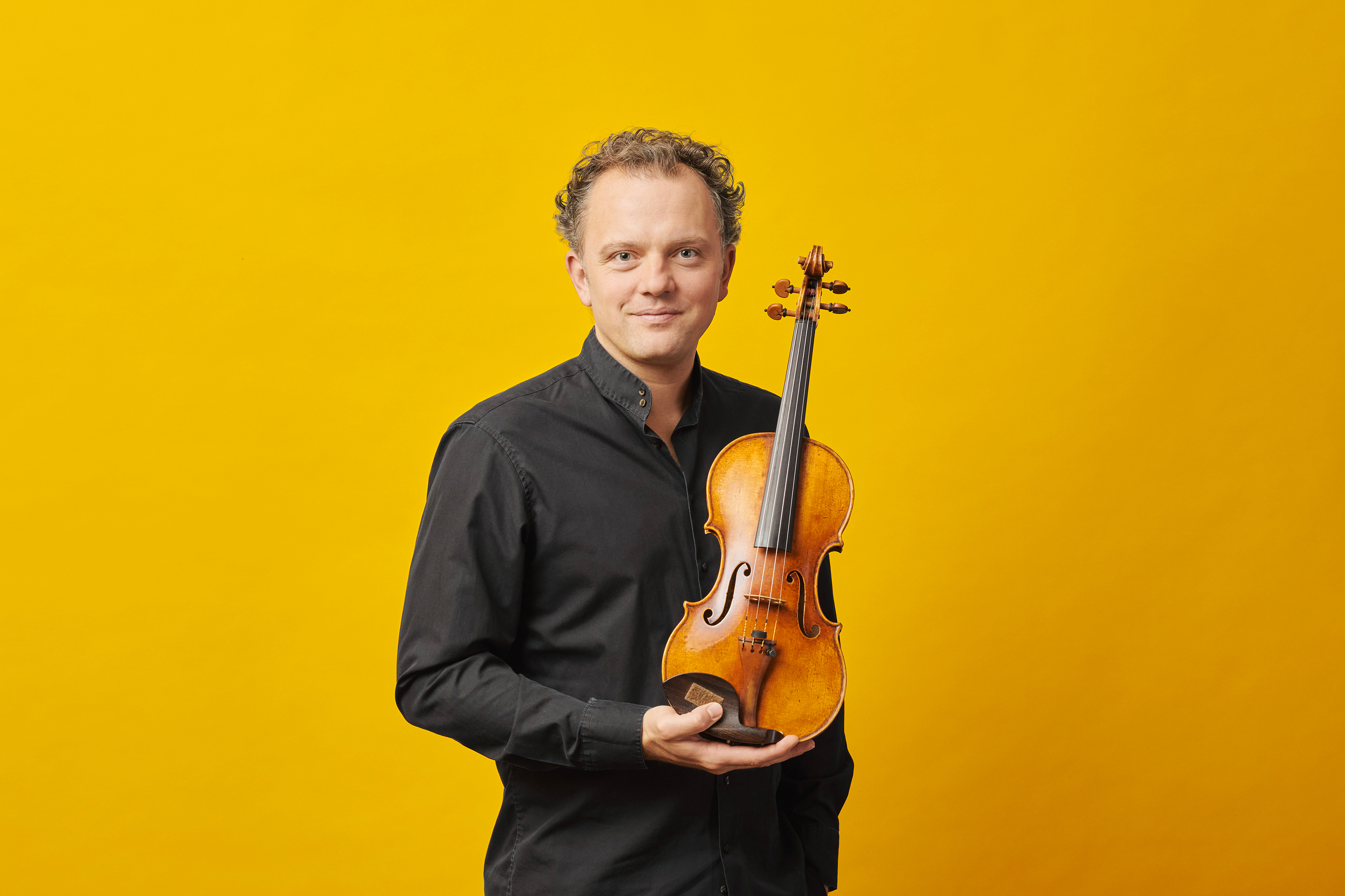 Portrait against a yellow background. Philipp Bohnen holds his violin in his hand.