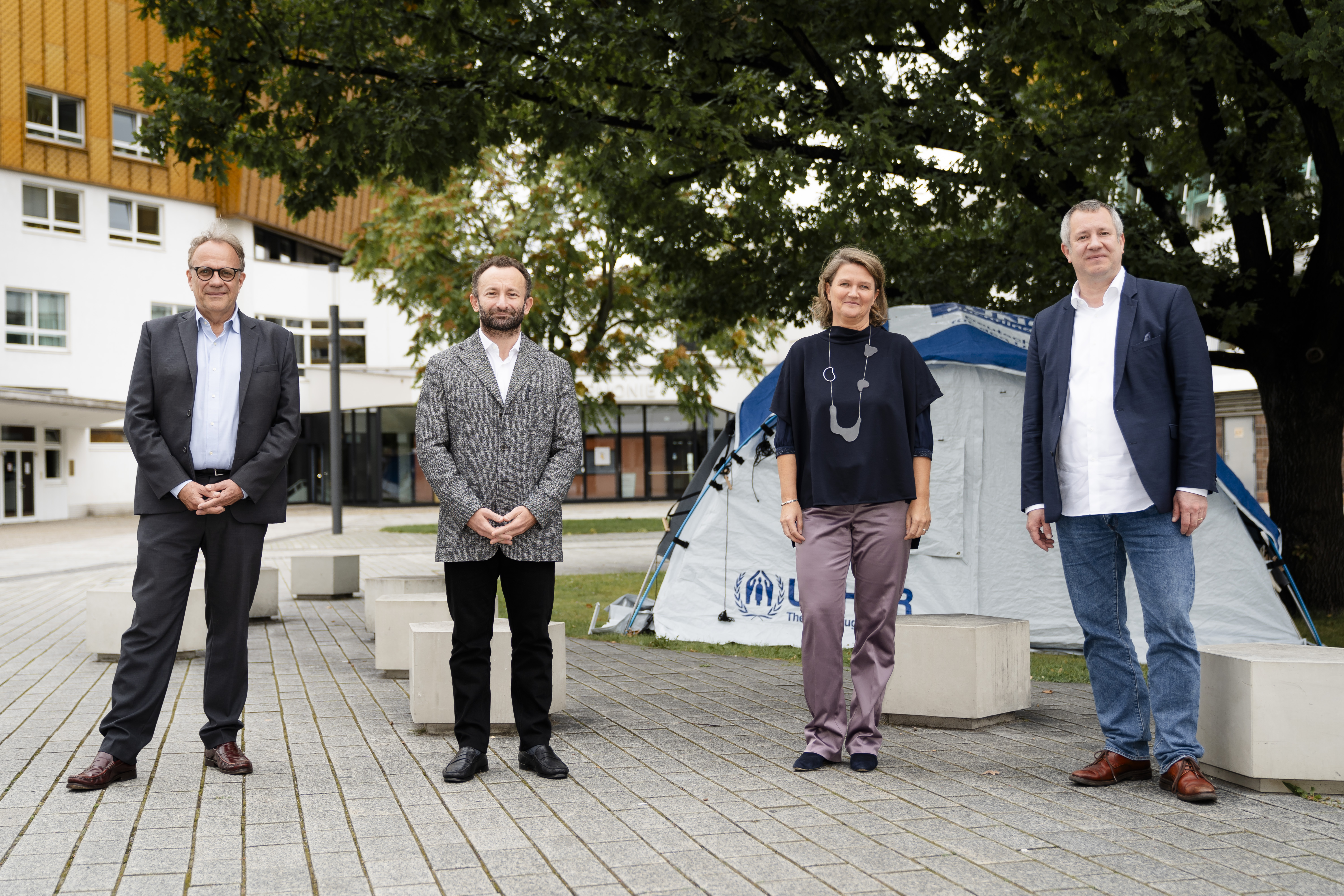 Peter Ruhenstroth-Bauer, Kirill Petrenko, Andrea Zietzschmann and Stefan Dohr standing in front of the Philharmonie. In the back there's a refugee tent of the UNHCR.