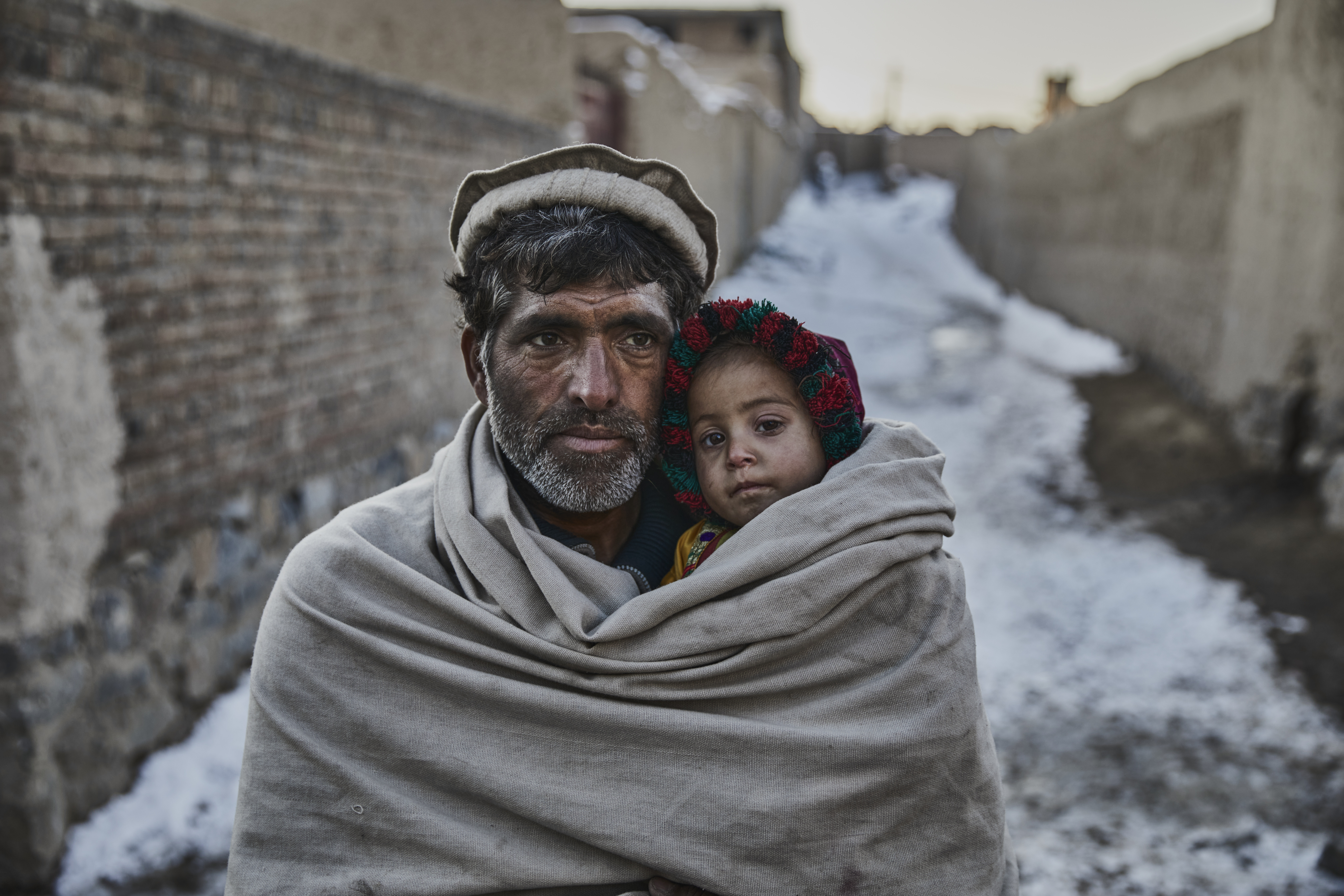 A man carries a small child in a warm blanket through a snow-covered alley. Both have serious faces; the child is wearing a colourful cap. The surroundings appear cold and plain, with stone walls and snow in the background.